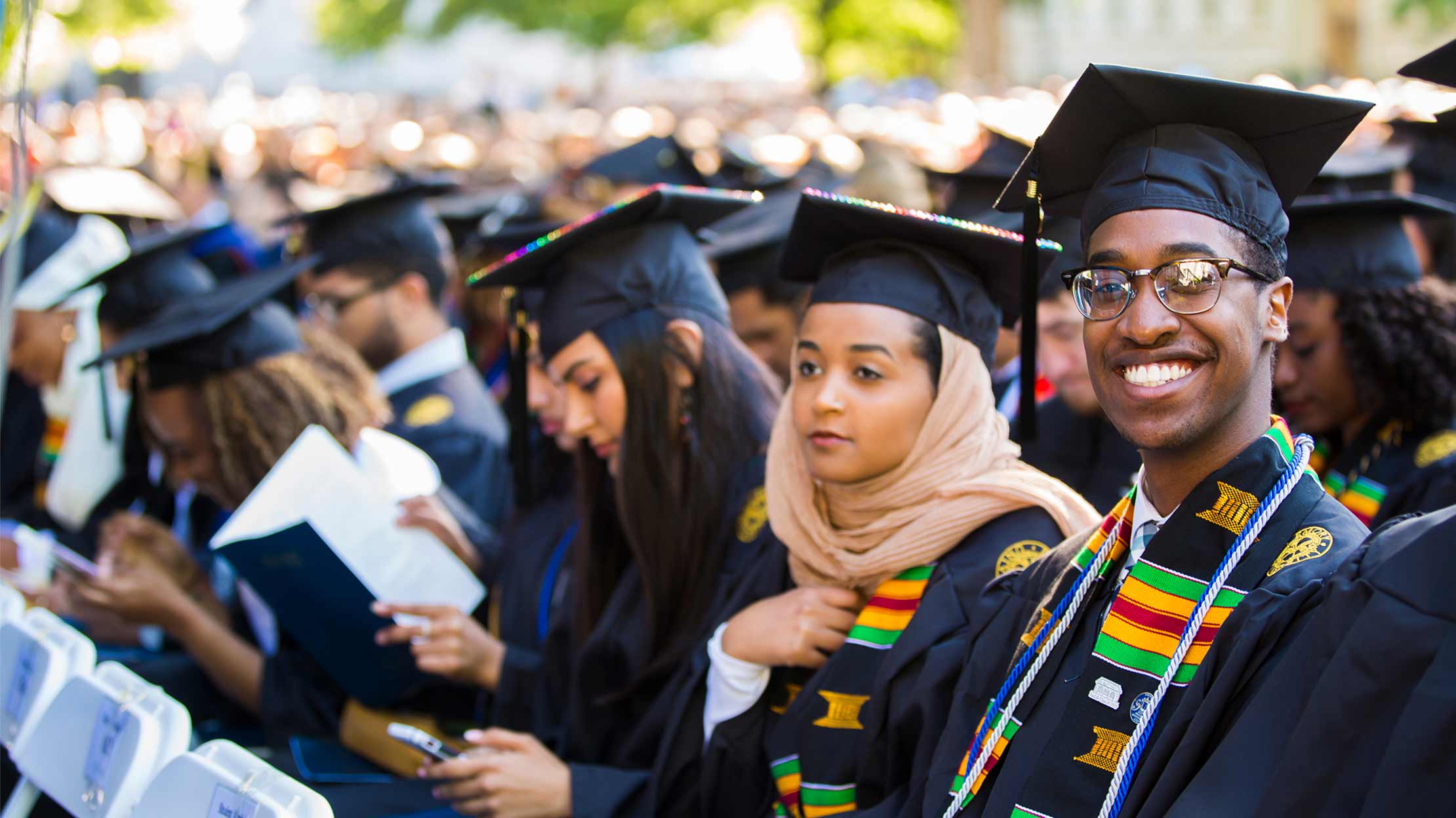 graduates sitting at commencement, including a young Black man smiling directly at the camera